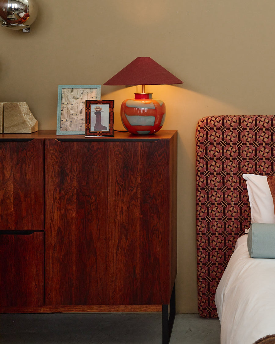 A styled bedroom corner featuring a bed with a patterned reddish-brown headboard, white bedding, and decorative pillows, next to a dark wood nightstand with a lit red lamp and wall-mounted metallic spheres.