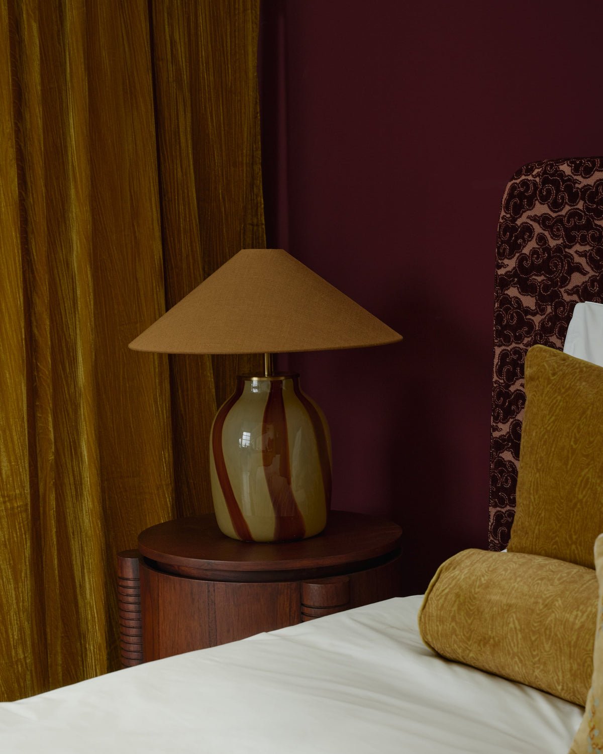 A cozy bedroom scene showing a bed with white bedding, mustard yellow velvet pillows, and a patterned headboard against a deep burgundy wall, with a matching bedside table lamp and gold curtains.