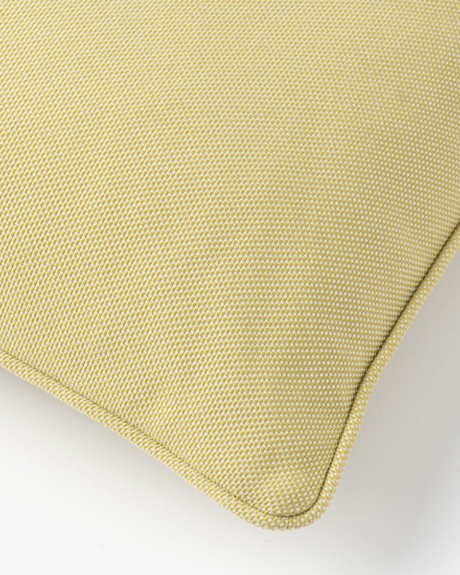 A close-up of a textured throw pillow in a light yellow-green or sage yellow fabric, showing the corner and stitched edge against a plain light gray background.