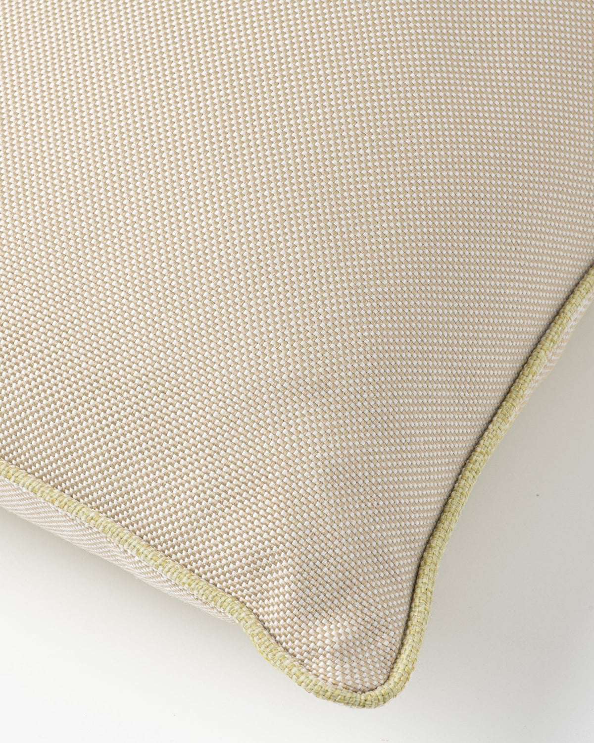 Close-up of a woven throw pillow corner showing a textured beige fabric and a light olive green piped edge on a white background.