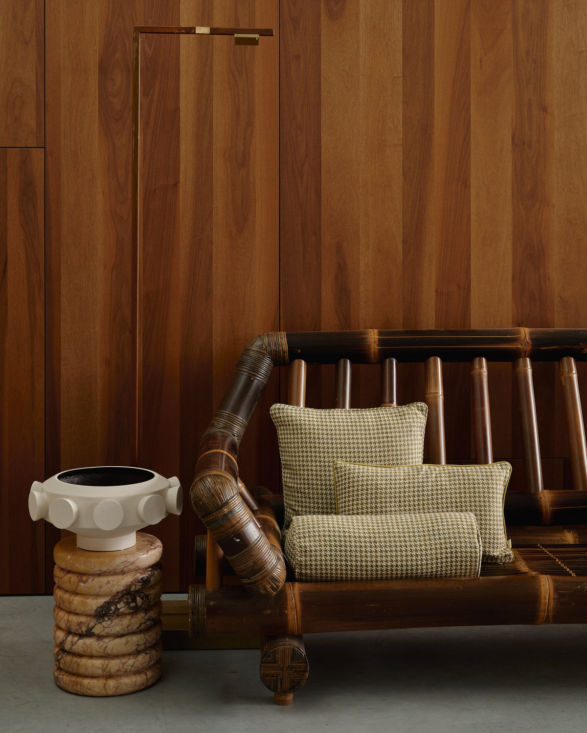 A rustic bamboo lounge chair with dark brown wooden slats and two beige-and-brown checkered pillows, set against a warm wood-paneled wall with a small carved stone side table beside it.