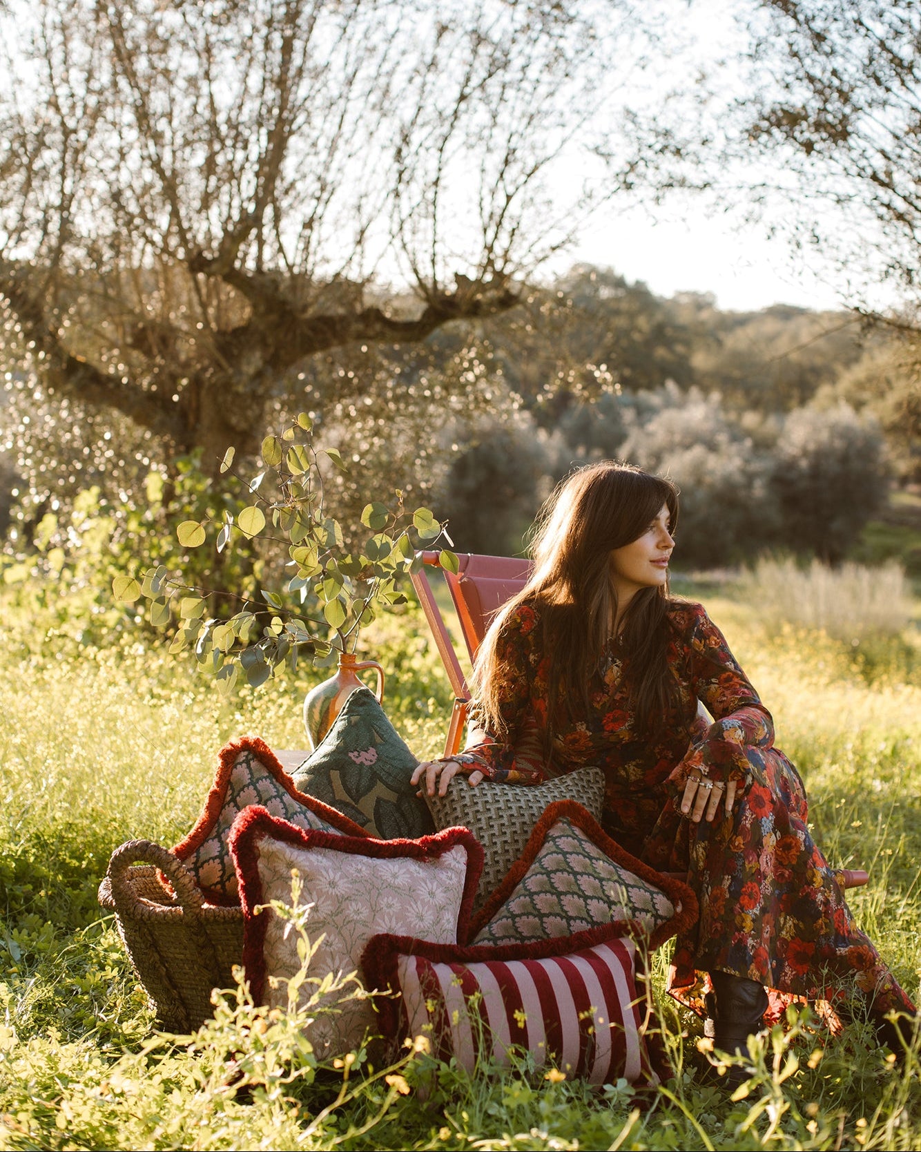 A woman sits outdoors in a grassy field near trees, wearing a long-sleeve bohemian-style floral dress. She is surrounded by patterned cushions and a basket in a sunlit outdoor setting.