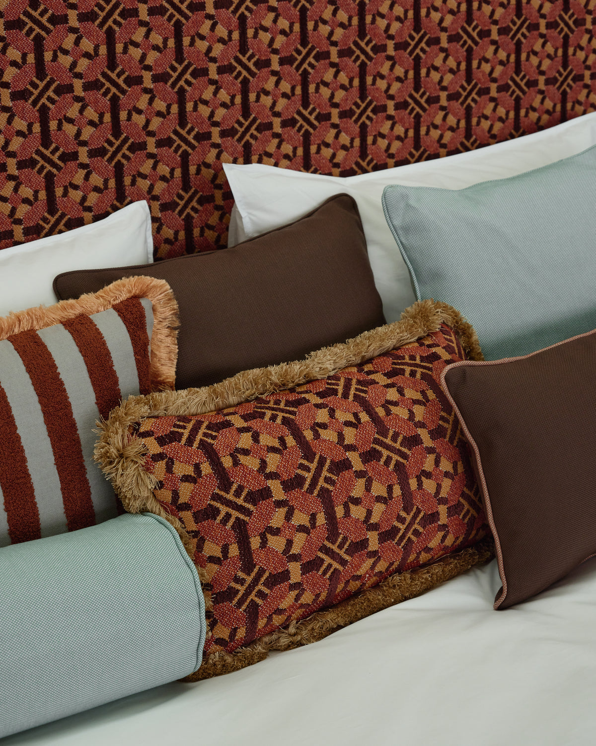 A styled collection of decorative throw pillows arranged on a bed with white linens and a patterned headboard. The pillows include solid dark brown, light blue, rust and beige geometric patterns with fringe trim, and a rust and beige striped pillow with a brown fringe.