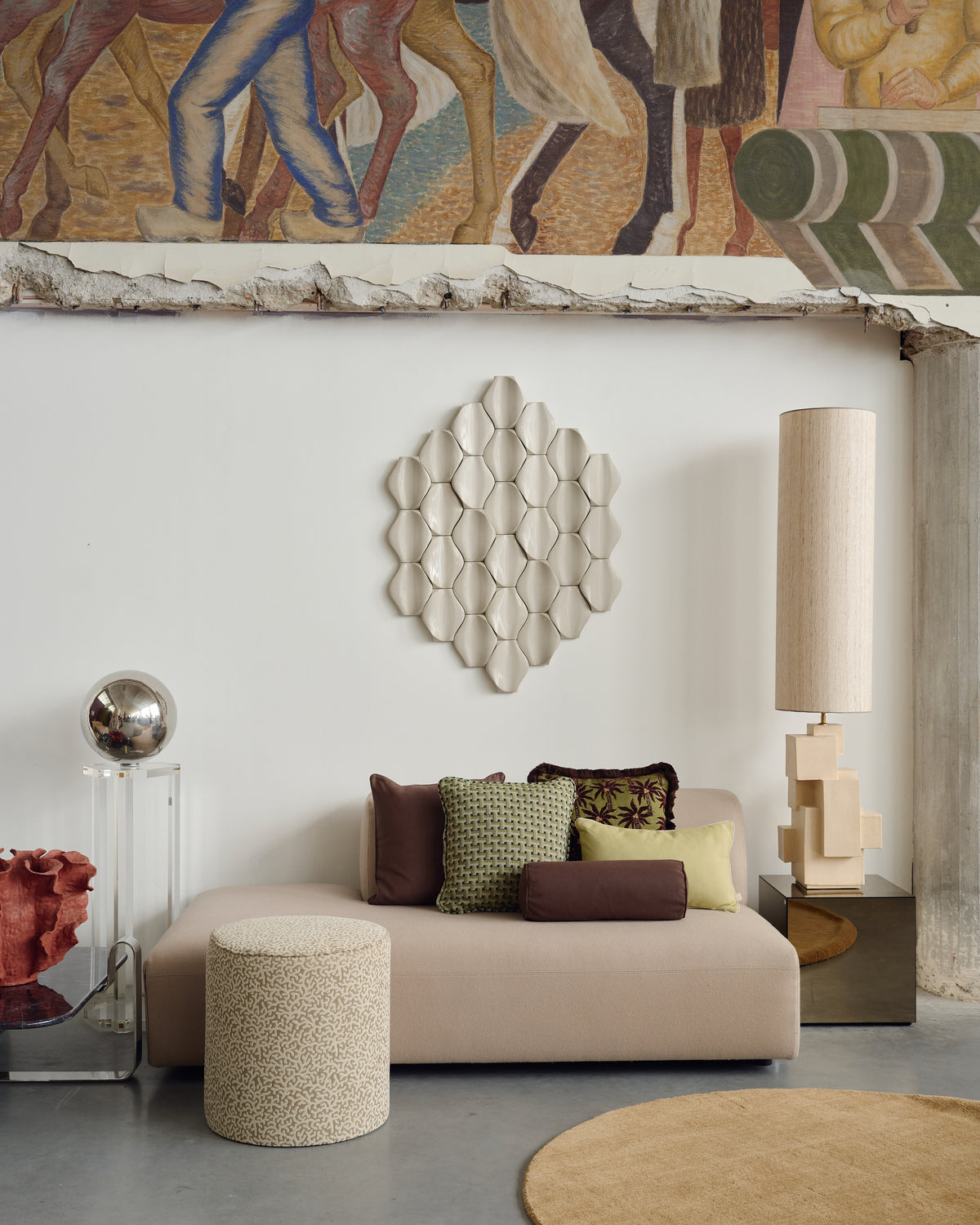A beige upholstered sofa in a styled living room setting, decorated with brown, green, and yellow accent pillows. A matching textured beige cylindrical ottoman sits in front of the sofa.