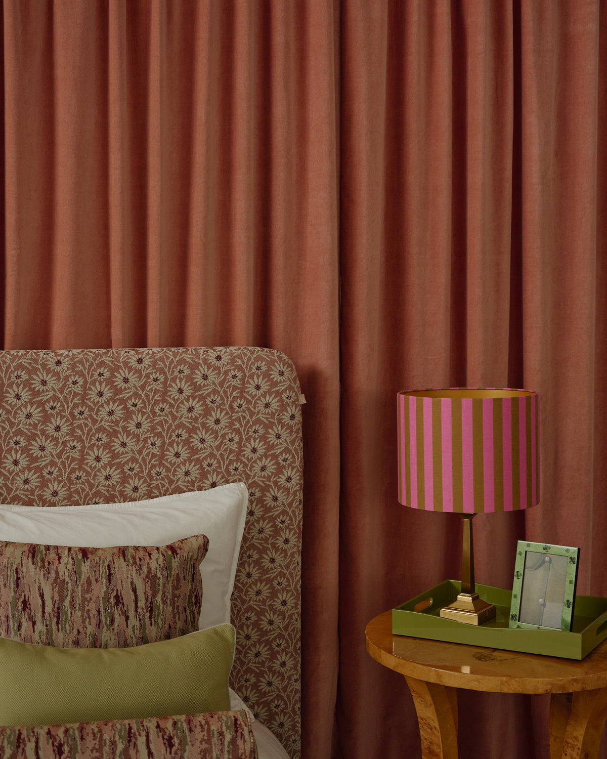 A styled bedroom corner featuring rust-brown velvet curtains, a patterned upholstered headboard, pillows, a striped pink and gold table lamp, and a small wooden side table.