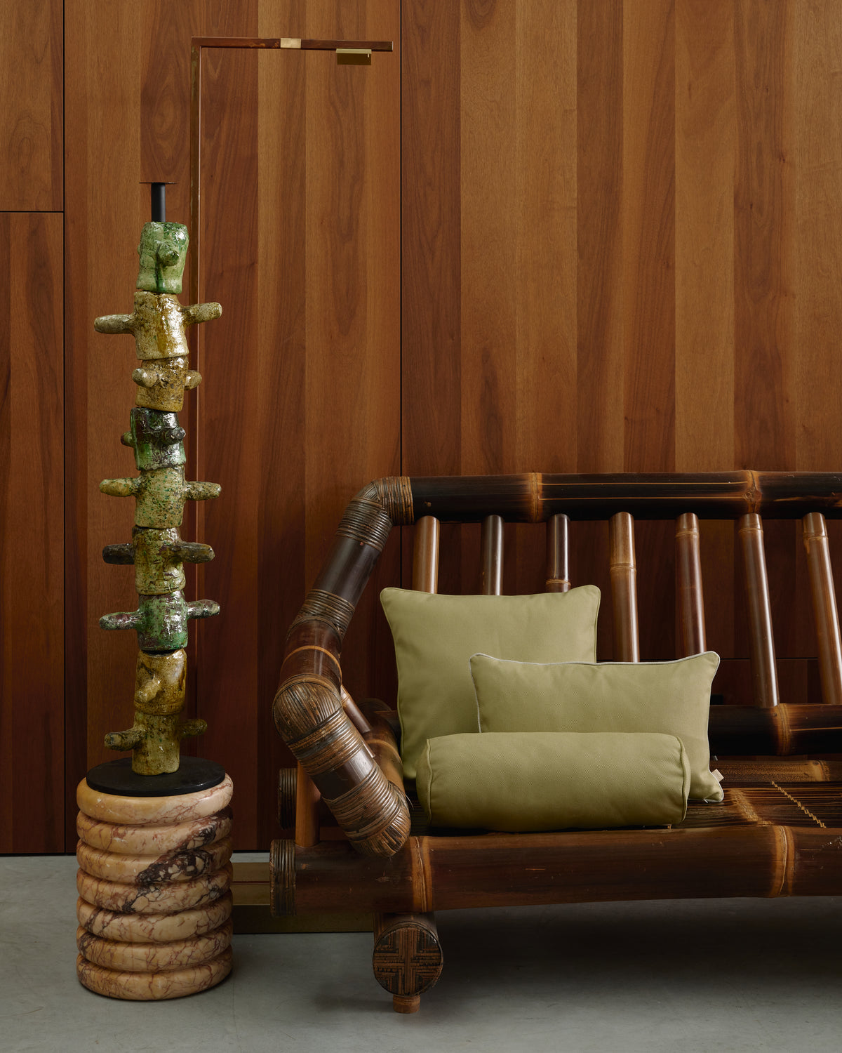 A styled interior scene with a dark wood rattan sofa adorned with olive green pillows, set against a matching wood-paneled wall. To the left stands a tall, decorative ceramic totem sculpture on a carved stone base.