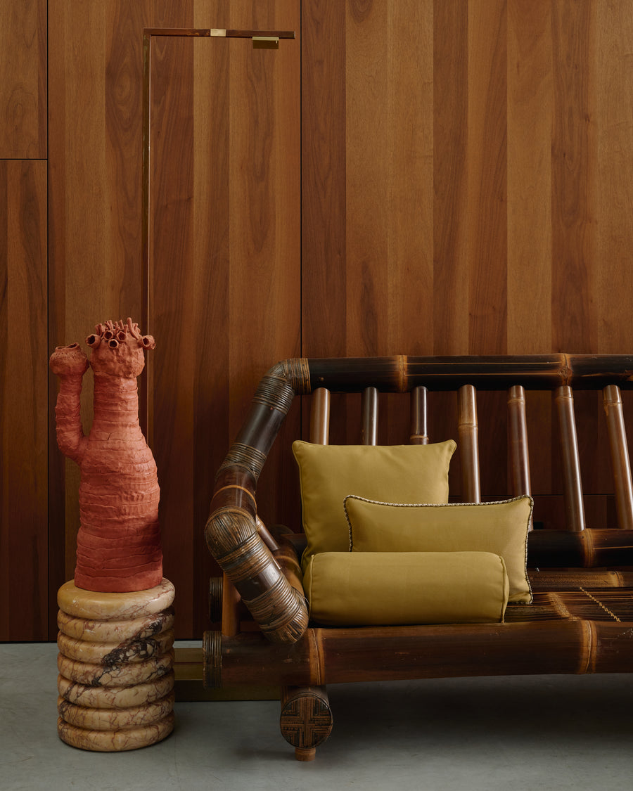 A rustic wooden bench with dark brown slatted backrest and seat, styled with two mustard yellow cushions, set against a matching wood-paneled wall. A terracotta-colored sculptural plant holder sits on a stone base to the left.