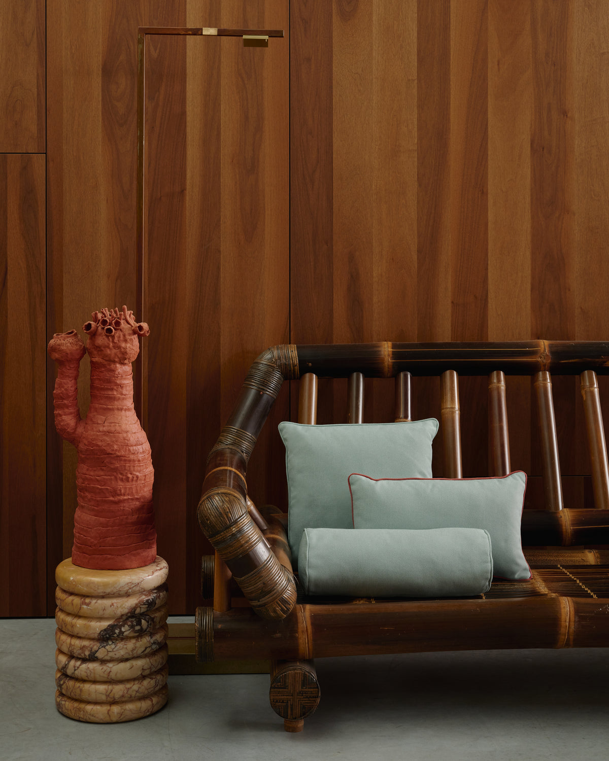 A modern bamboo lounge chair with light blue cushions, set against a dark wood-paneled wall next to a terracotta-colored sculptural vase on a stone pedestal.