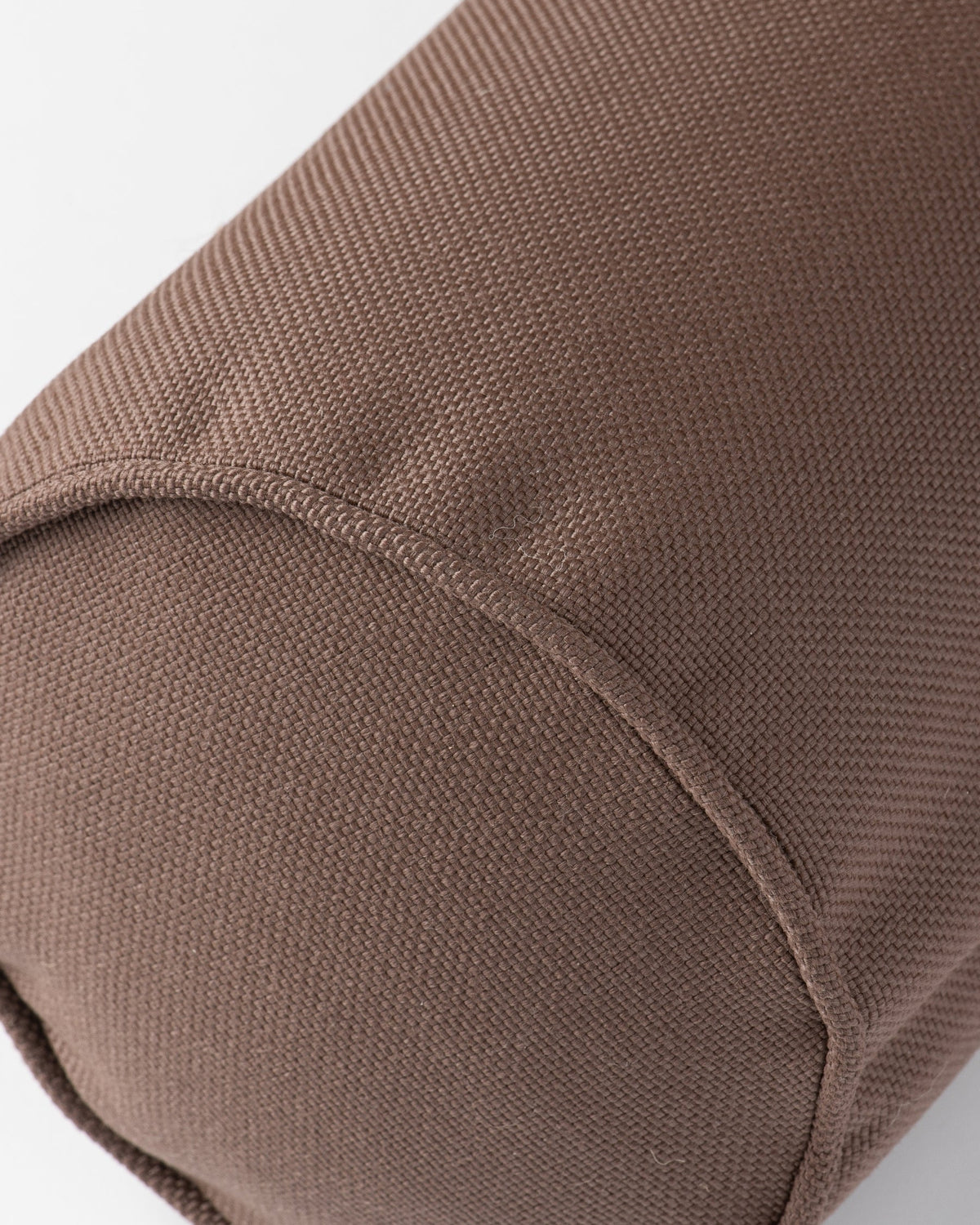 Close-up of a brown woven fabric cylindrical cushion or bolster with a curved seam, shown against a plain light gray background.