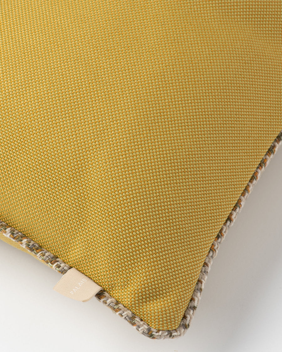 Close-up of a mustard yellow woven cushion cover with a textured beige and gray braided trim along the edge, showing a small fabric tag.