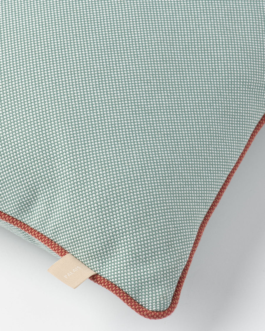 Close-up of a square throw pillow in a sage green and white micro-check pattern with rust-red piping trim and a small beige fabric brand tag.
